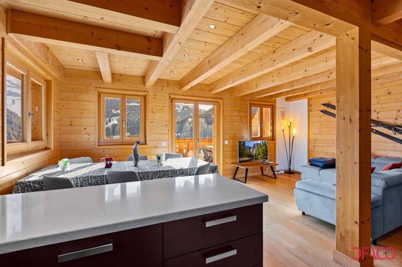 Spacious open-plan living area with wooden walls and ceiling, large windows offering scenic mountain views, modern kitchen island with granite countertop, comfortable sofas, and a TV stand