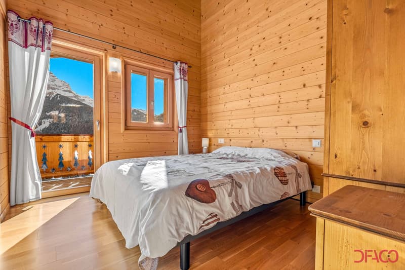 Cozy wooden bedroom with large windows offering a scenic mountain view, wooden walls and floors, a double bed with patterned bedding, and a small side table.