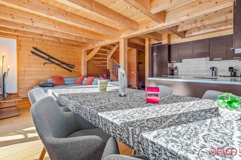 Spacious open-plan living area with wooden ceiling and walls, large sofa, dining table, kitchen area with modern appliances, and a fireplace visible on the wall.