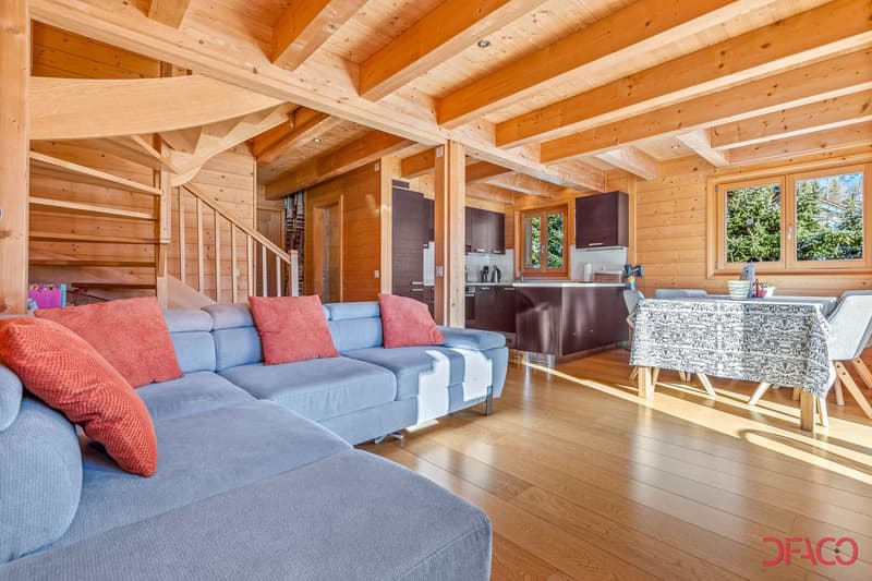 Spacious open-plan living area with wooden beamed ceiling, large windows, hardwood floors, comfortable blue and red sofas, and a kitchen area with dark cabinets and appliances