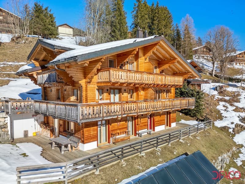 2-story wooden chalet-style house with a large balcony, surrounded by snow-covered trees and hills in a mountainous winter landscape