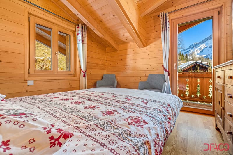 Cozy wooden bedroom with large windows offering a scenic mountain view, a patterned bedspread, and two gray armchairs.