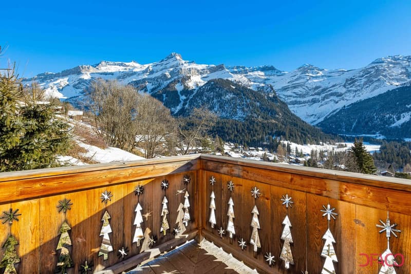 Wooden balcony with decorative snowflake and tree ornaments, overlooking a snowy mountain landscape with a village in the foreground