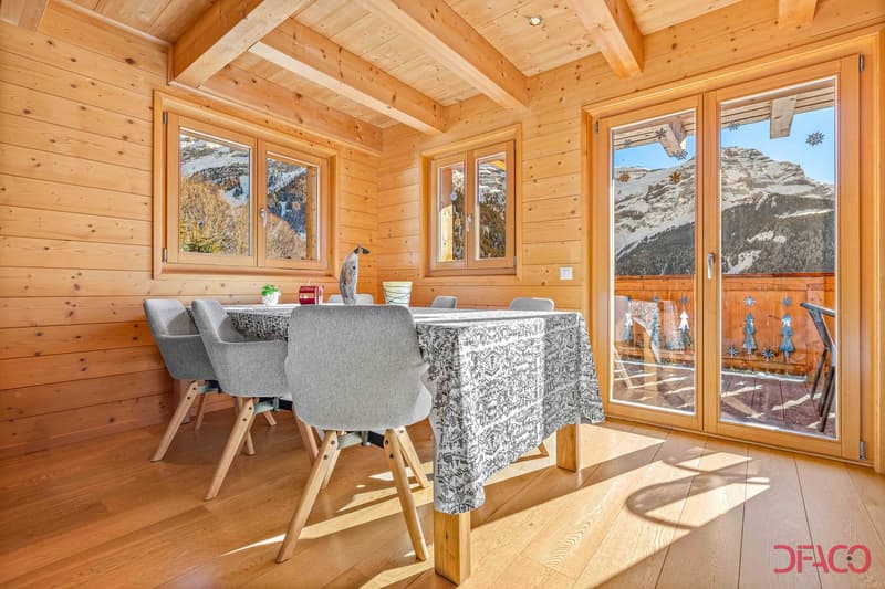 Spacious dining area with wooden walls and ceiling, large windows offering a scenic mountain view, wooden dining table with patterned tablecloth and gray chairs, decorative elements on the table