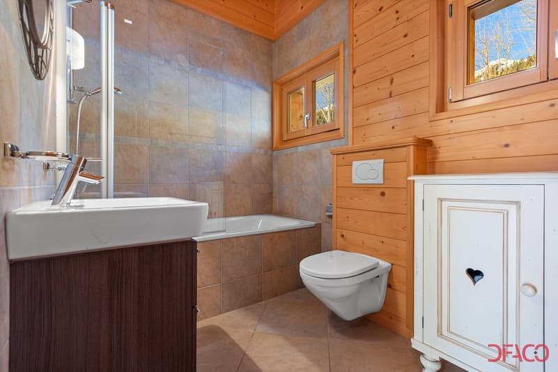 Spacious bathroom with tile flooring, wooden walls, large vanity sink, and a toilet. The bathroom has a large window providing natural light and a view of the outdoors.