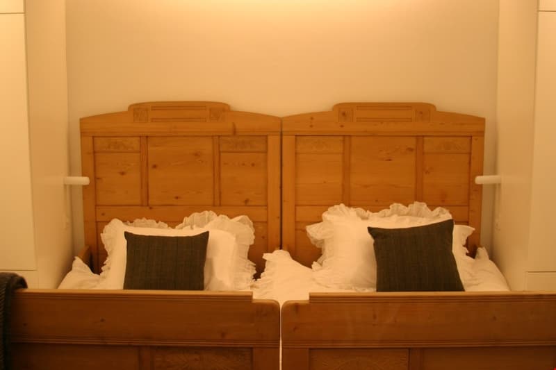 The image shows a bedroom with two wooden headboards, each with drawers and cabinets. The bed is made with white bedding and black accent pillows.