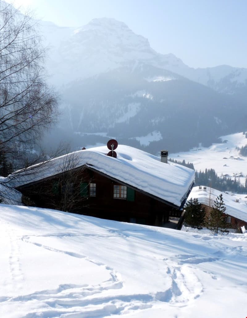 Wooden chalet-style house with a snow-covered roof, surrounded by snowy mountains and trees in the background.