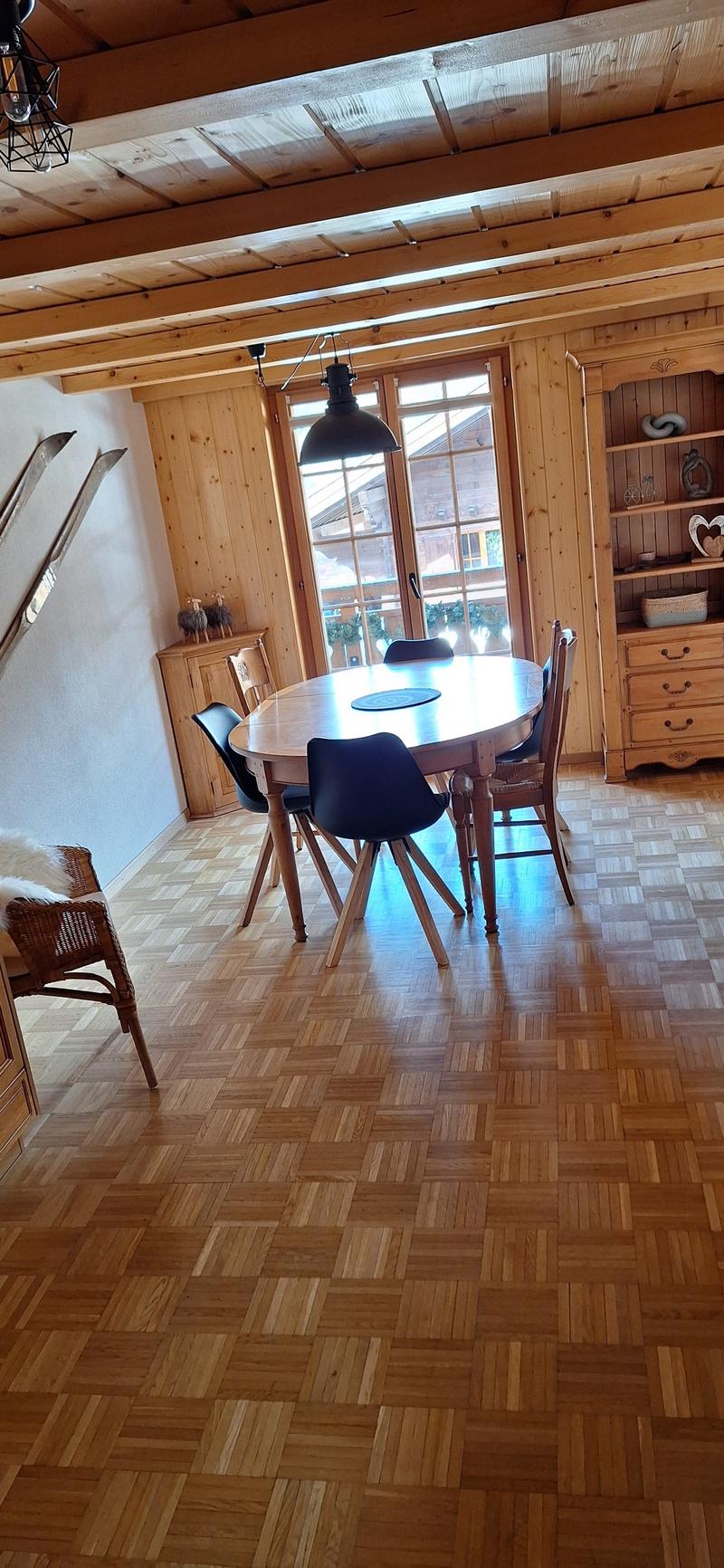 Wooden dining table with chairs, wooden shelves, wooden beams on the ceiling, large window providing natural light, and a fireplace visible in the room.