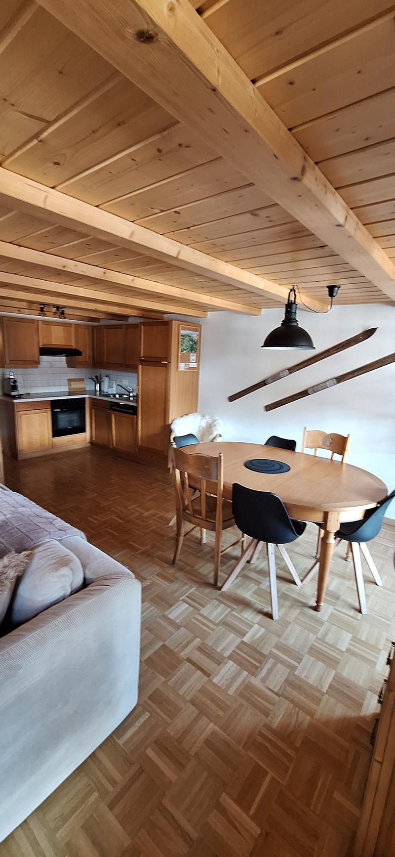 The image shows an open-plan living space with a kitchen, dining area, and living room. The space features wooden beams on the ceiling, a wooden dining table with chairs, a sofa, and a kitchen with wooden cabinets and a dishwasher.