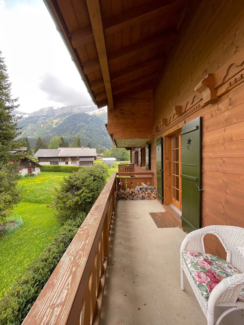 Wooden cabin with a covered balcony/terrace, overlooking a scenic mountain landscape with forests and buildings in the distance. The balcony has a wicker chair and firewood stacked nearby.