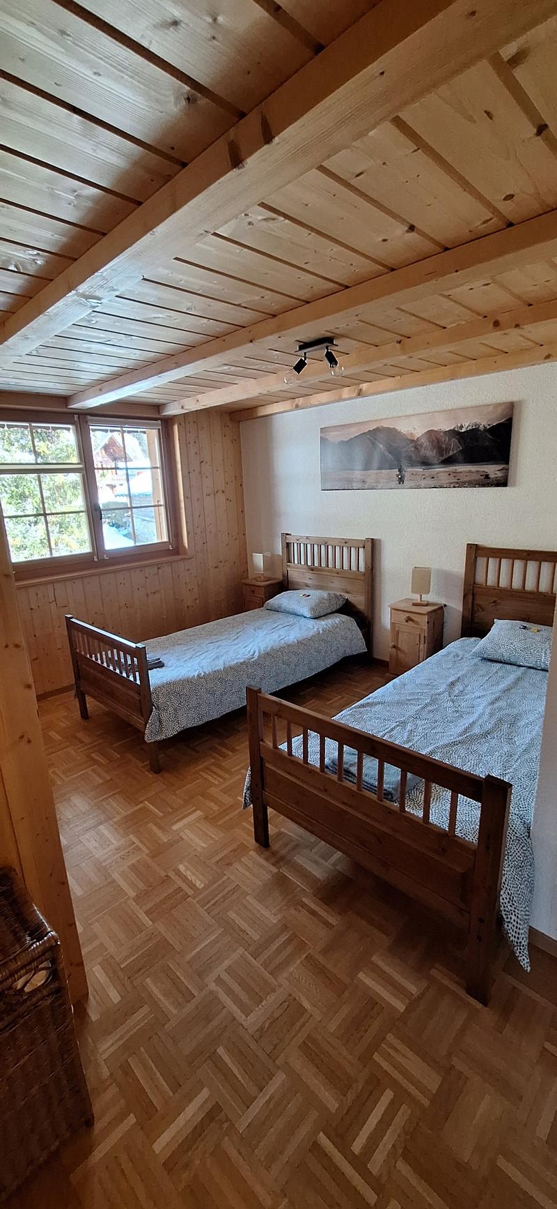 Cozy wooden bedroom with two twin beds, wooden ceiling beams, and a large window overlooking a scenic landscape. The room has a warm, rustic feel with wooden furniture and parquet flooring.