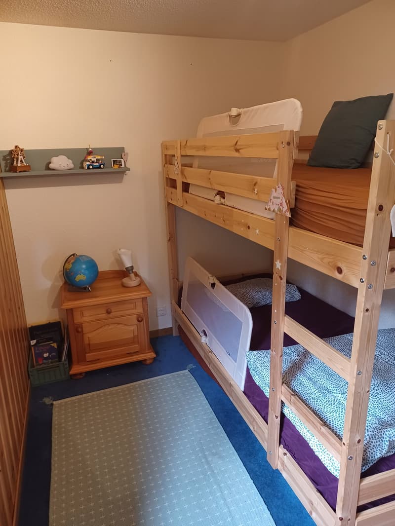 The image shows a bedroom with a wooden bunk bed, a wooden nightstand, and various toys and decorations on the shelves. The room has a blue and gray color scheme, with a patterned rug on the floor.