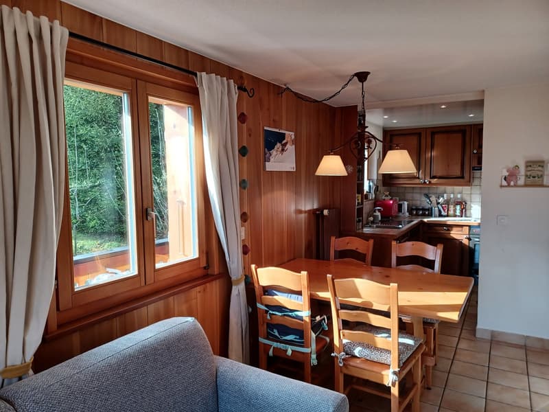 The image shows a cozy kitchen and dining area with wooden paneled walls, large windows overlooking a garden, a dining table with chairs, and a kitchen counter with appliances. The room has a warm, rustic feel with decorative elements like wall hangings.