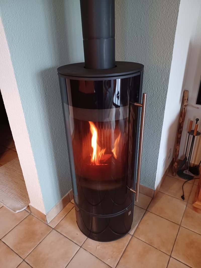 A modern, cylindrical wood-burning fireplace with a glass door, displaying a warm, flickering fire inside.