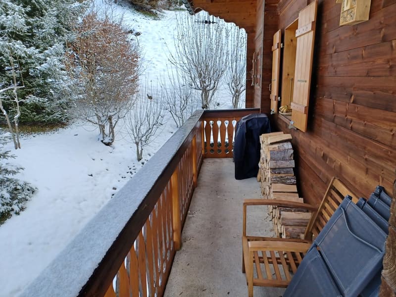 Wooden balcony with railing, overlooking a snowy landscape with trees and a wooden cabin in the background