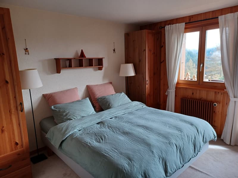 Cozy bedroom with wooden walls and furniture, large window with a view, queen-size bed with blue and pink bedding, wall shelves, and a lamp