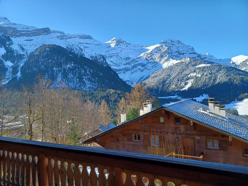 The image shows a scenic mountain landscape with snow-capped peaks, a wooden chalet-style building with a balcony, and a forested area in the foreground.