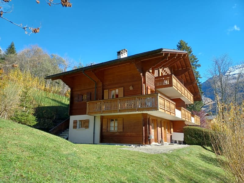 2-story wooden chalet-style house with a balcony, surrounded by trees and a grassy lawn in a mountainous setting