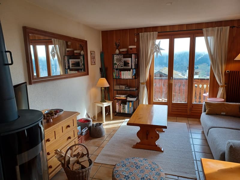 Cozy living room with wooden paneled walls, large windows overlooking a balcony, a fireplace, bookshelves, and a wooden dining table