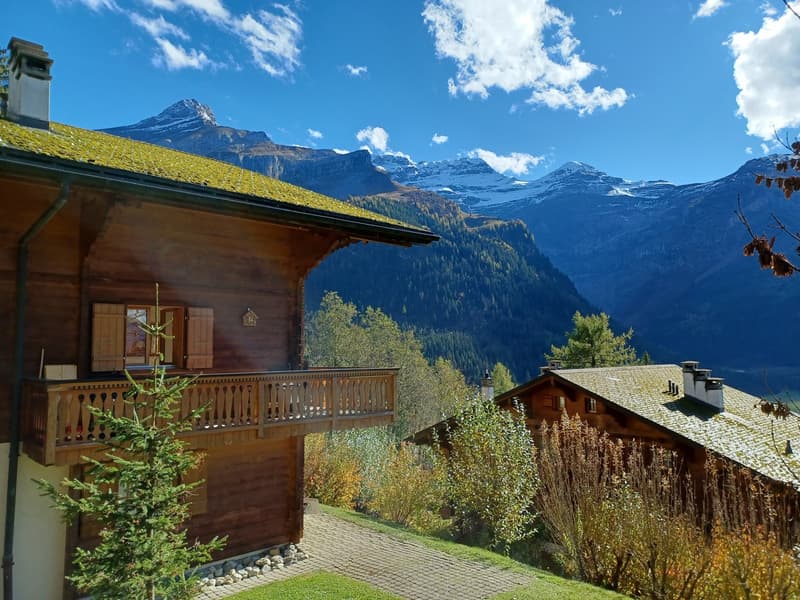 Wooden chalet-style house with a balcony, surrounded by mountains, forests, and a grassy yard