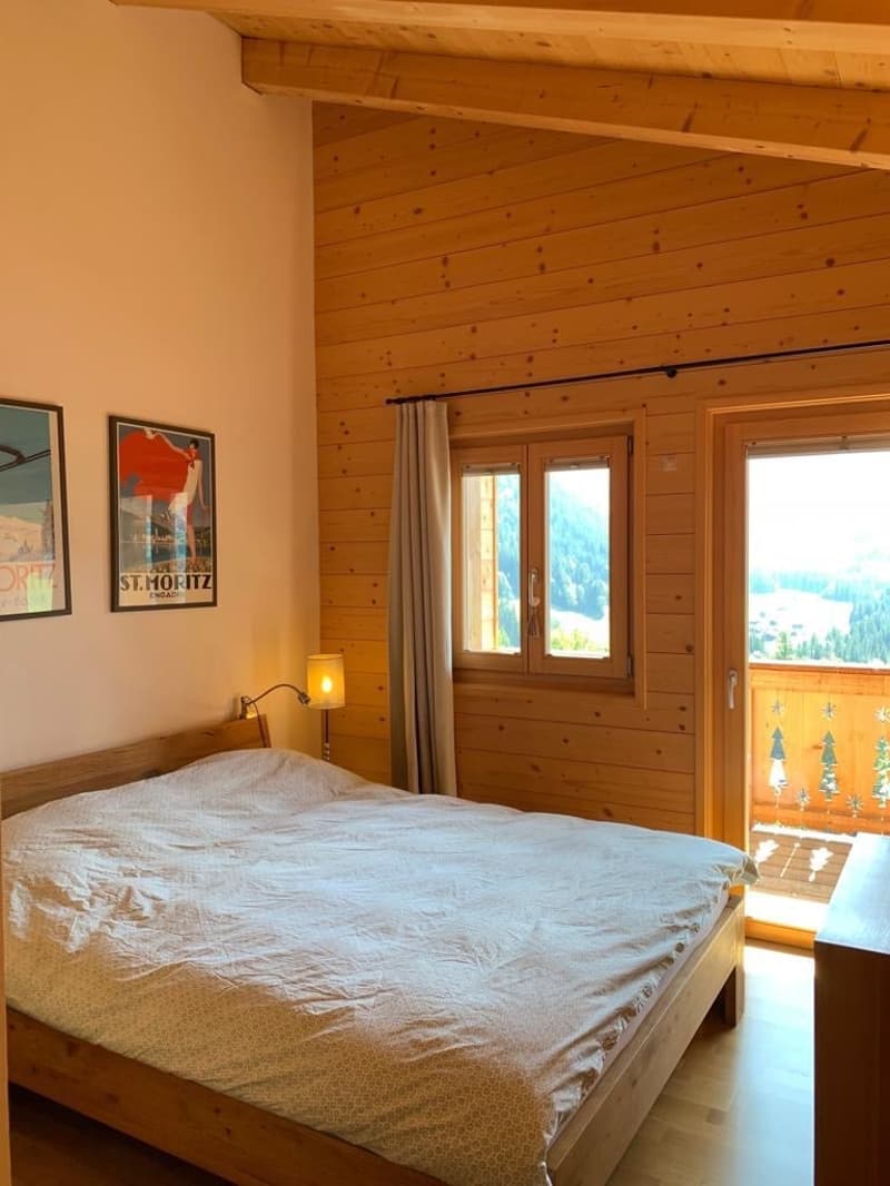 Cozy bedroom with wooden walls and ceiling, large windows overlooking a balcony or terrace, bed with bedding, and framed artwork on the walls.