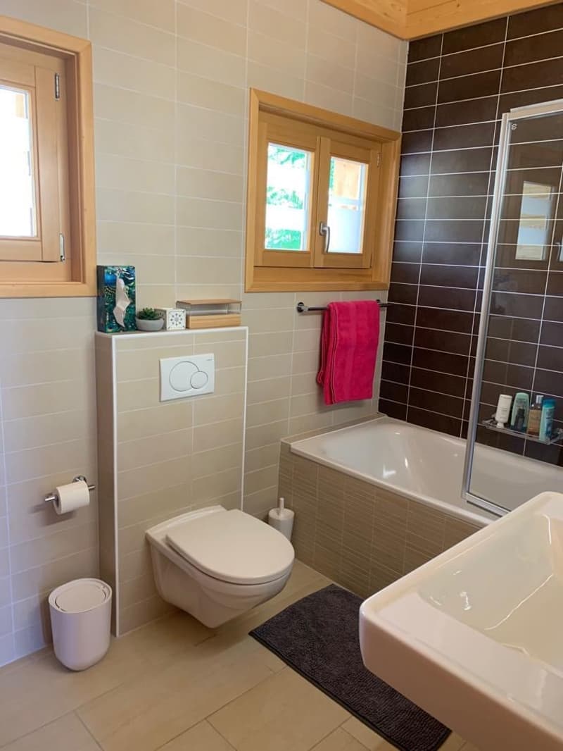 The image shows a modern bathroom with a toilet, bathtub, and shower. The walls are tiled in a light and dark color scheme, and there are wooden window frames. The bathroom also includes a towel rack and a small shelf unit.