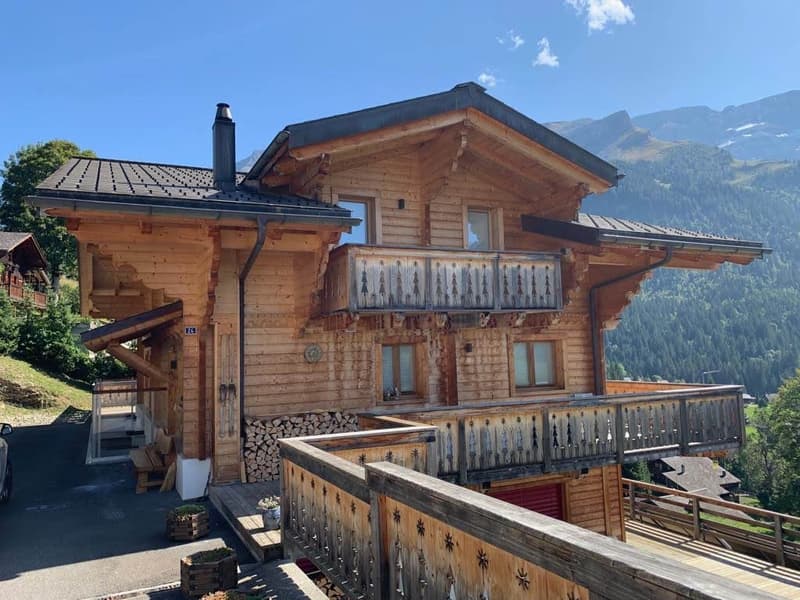 2-story wooden chalet-style house with a balcony, wooden railings, and a chimney. The house is surrounded by mountains and forests, creating a scenic alpine setting.