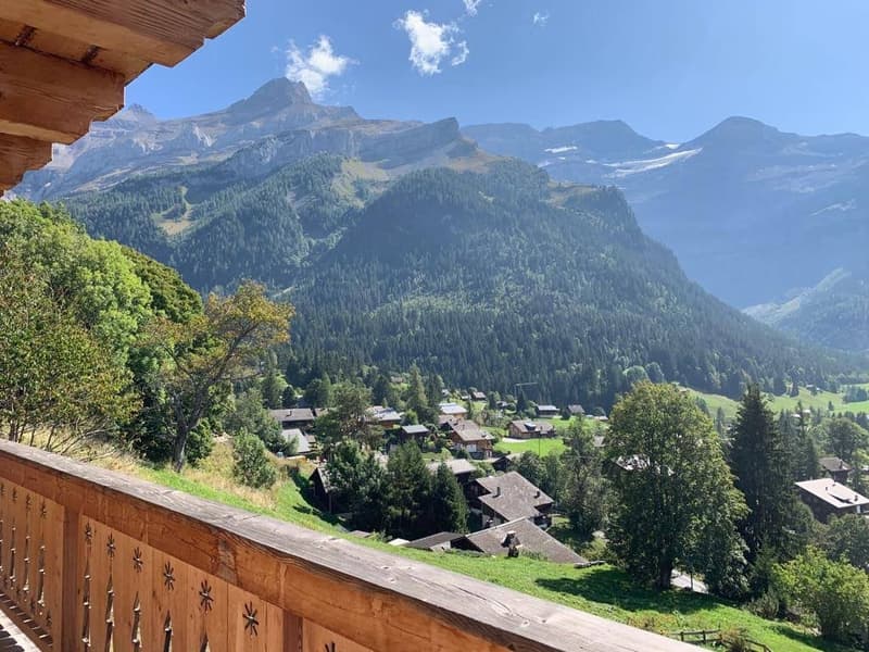 The image shows a picturesque mountain village nestled in a lush, forested valley. The village is surrounded by towering, snow-capped peaks and dense evergreen forests. The wooden balcony or terrace in the foreground offers a panoramic view of the stunnin