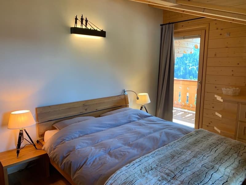 Cozy bedroom with wooden walls and furniture, large window with a balcony/terrace, bedside lamps, and a decorative light fixture above the bed.