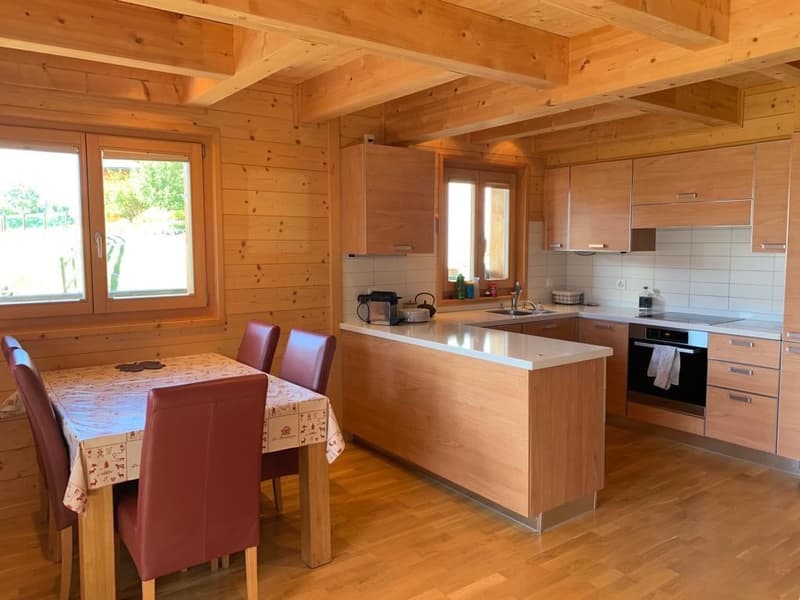 The image shows a spacious open-plan kitchen and dining area in a wooden cabin-style interior. The kitchen features modern appliances including a dishwasher, and the dining area has a wooden table and chairs. The room has large windows providing natural l