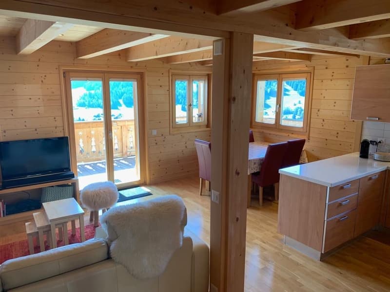 Cozy wooden cabin interior with large windows, open-plan living room and kitchen, comfortable seating, and access to a balcony or terrace with scenic mountain views.