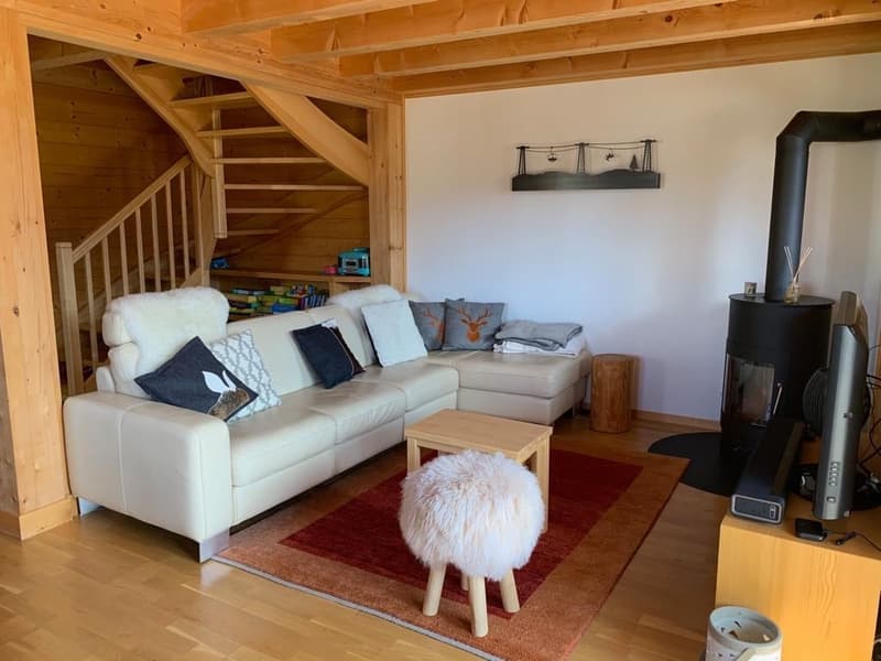 Cozy living room with wooden beams, white leather sofa, coffee table, and a fireplace. The room also has a staircase leading to an upper level.