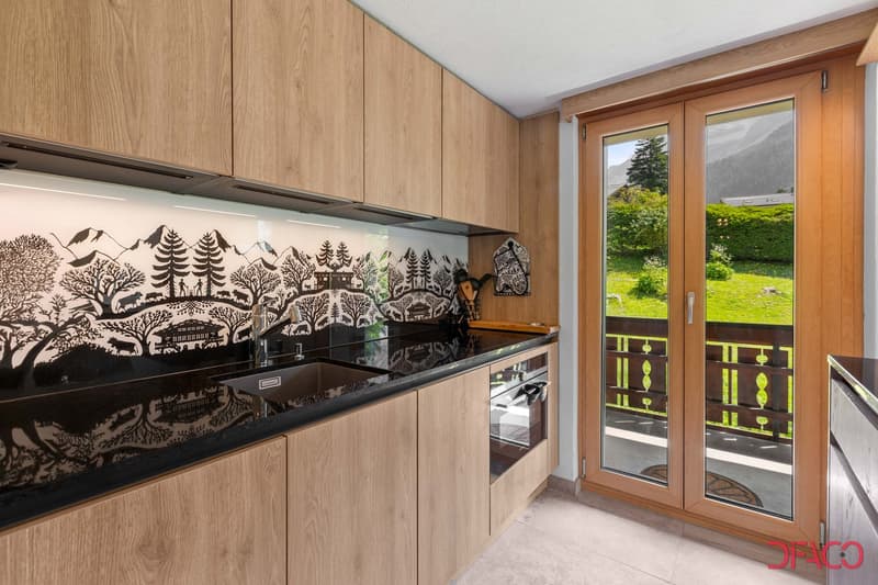 Wooden cabinets, black countertop, sink, faucet, large windows, wooden door leading to balcony, scenic mountain view, wooden floors, integrated oven