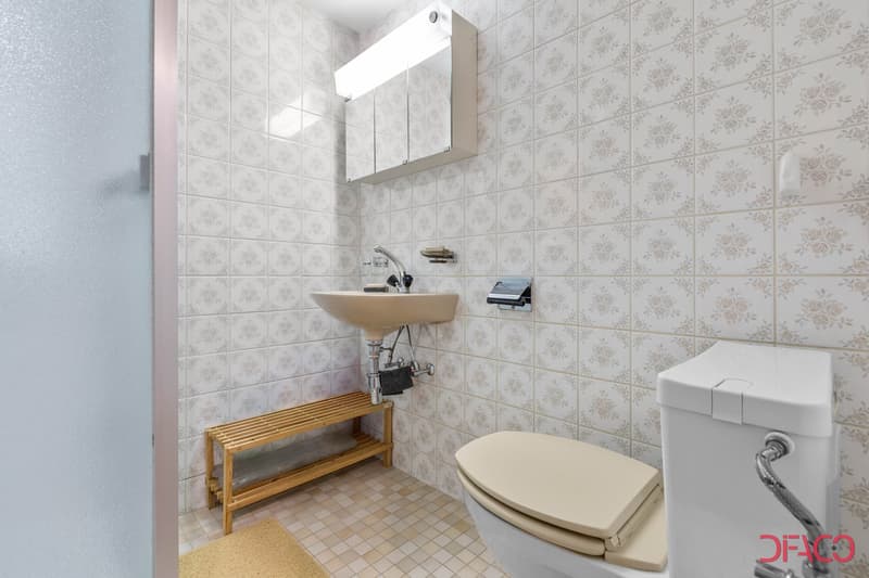 tiled walls, tiled floor, single vanity with sink, wooden bench, toilet, white shower curtain, mounted mirror, illuminated mirror above vanity