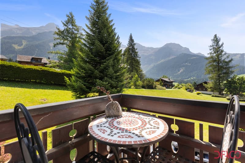 Balcony with round mosaic table, 2 chairs, decorative rock, wooden railing, fenced grass, trees, car, mountains in distance