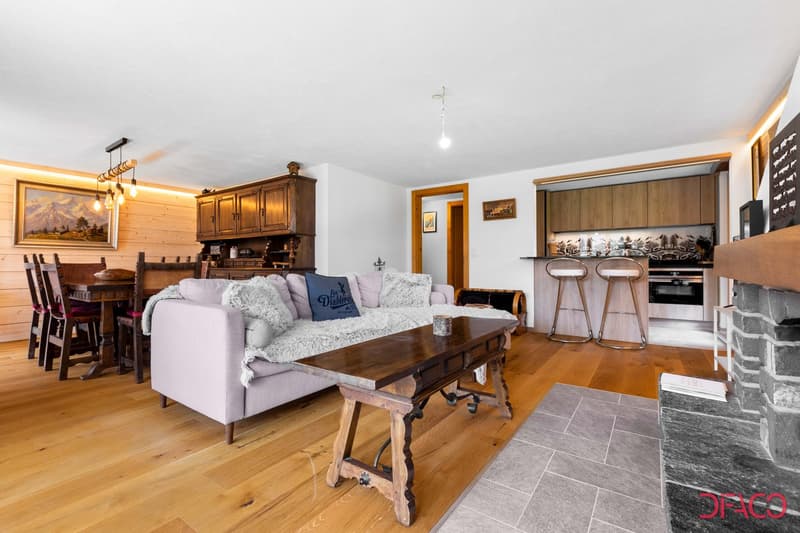 Light wooden floors, wooden dining table with chairs, large sofa with pillows, coffee table, kitchen bar with stools, stone fireplace, cabinets, two picture frames on wall