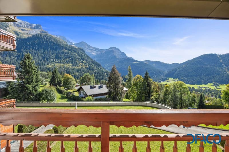 balcony with wooden railings overlooking mountains, trees, and green fields