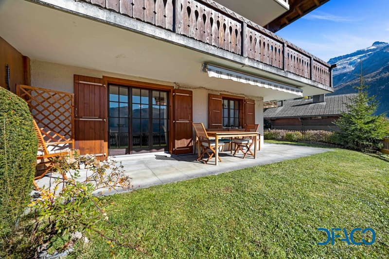 A modern apartment with large windows, a wooden terrace, a wooden table and chairs, and a grassy garden with bushes and a plant.