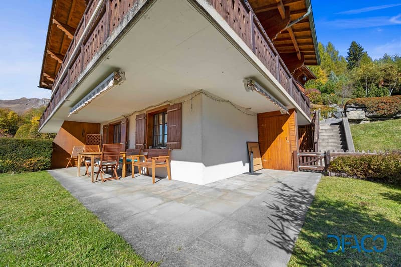 Alpine-style house with balcony, wooden floors, white and brown exterior, patio with table and chairs, stairs leading to upper level, garage, lush garden with hedges and shrubs, surrounded by nature