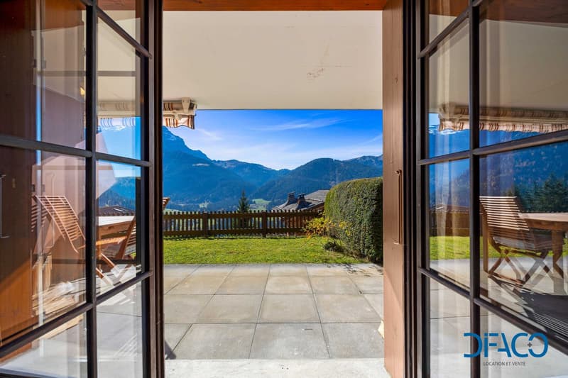 Glass doors opening to a patio with outdoor seating, manicured lawn, wooden fence, mountains view