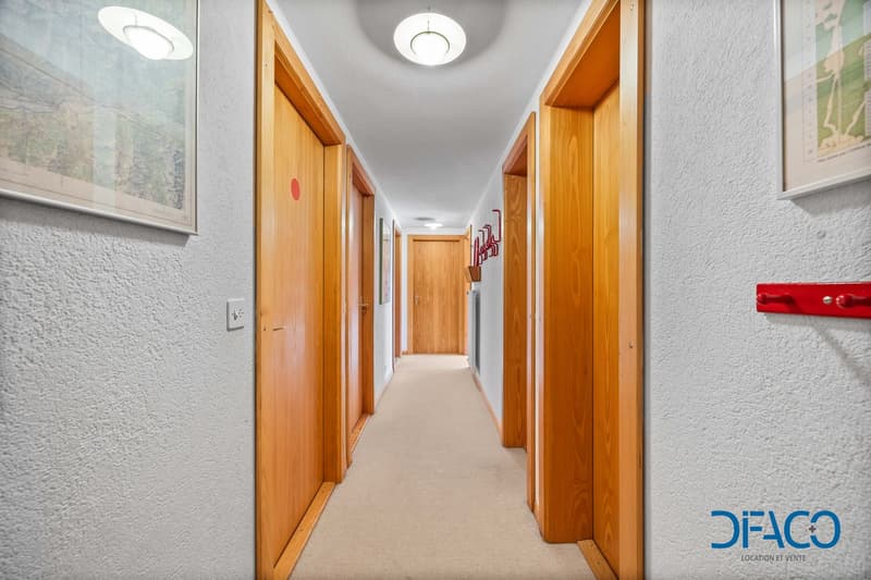 a narrow hallway with wooden doors and beige carpet flooring, white walls, and mounted pictures