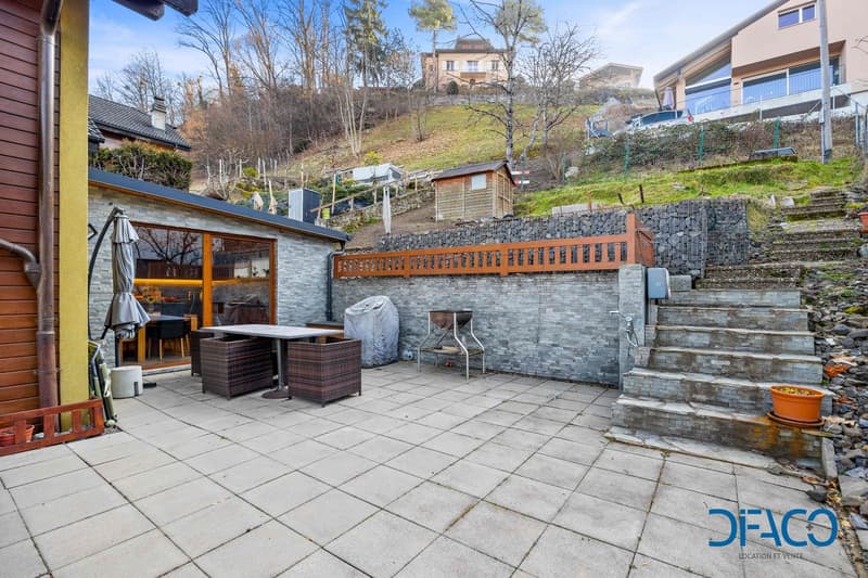 A sloped backyard with stone steps leading to a patio, a table and chairs, and a small shed. It has a stone wall with a small window and a stone patio.