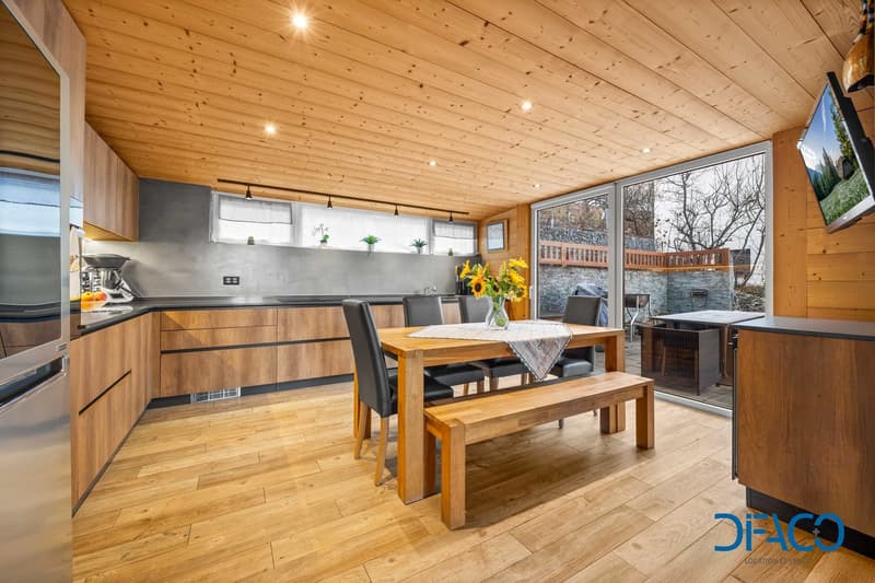 Modern kitchen with wooden flooring, wooden walls, kitchen island with dining table, wooden bench, sliding doors, windows, appliances, television.