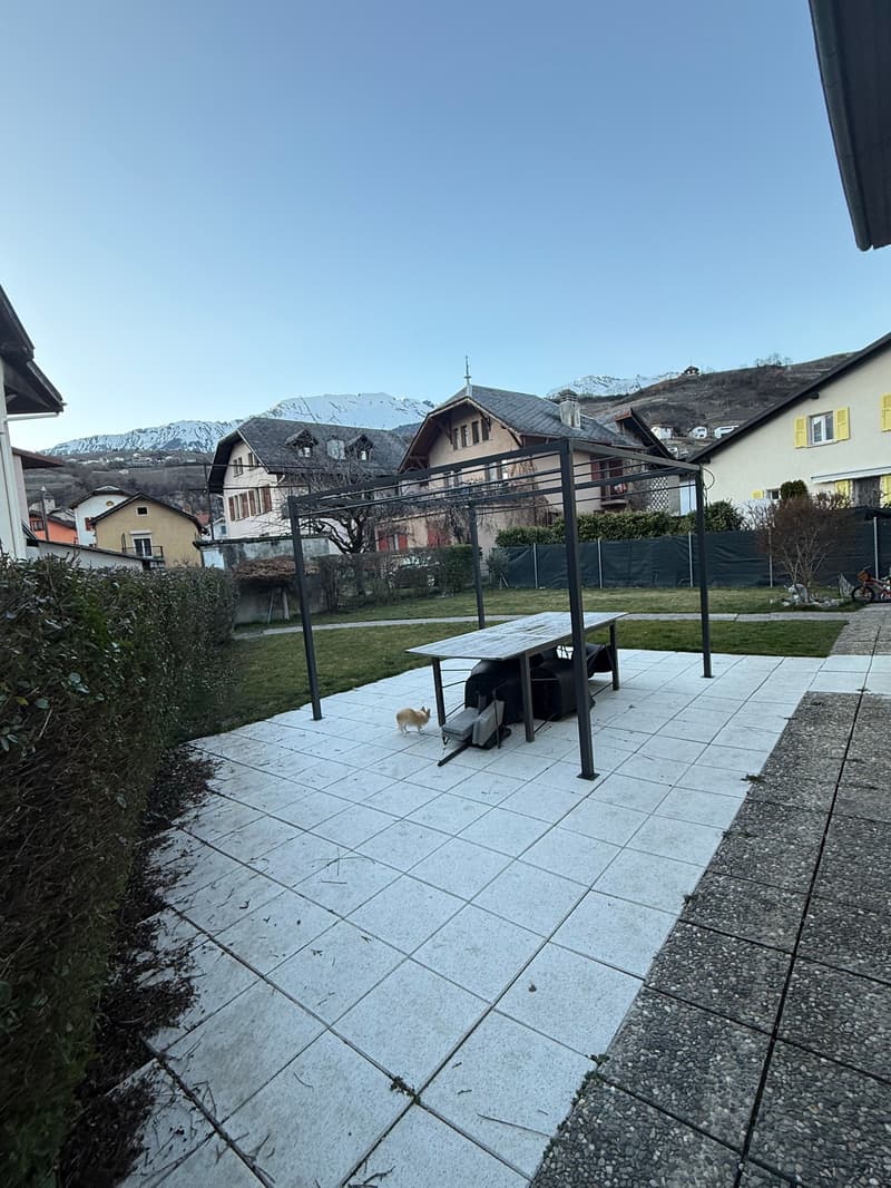 outdoor patio area with table and canopy, green lawn, bushes and trees, mountain view in the distance