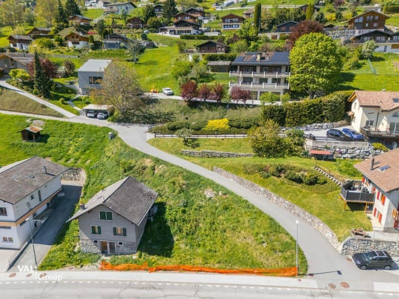 Multiple chalets and houses, situated on a hillside with a view, small roads, parking spots for the vehicles, greenery, and trees in the area