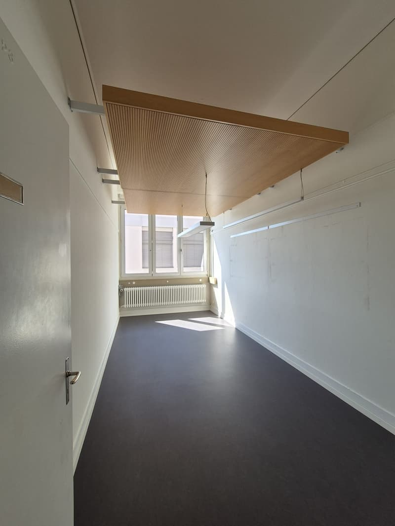 An empty room with wooden ceiling, white walls, and black flooring. It has a window with a radiator underneath and fluorescent lights hanging from the ceiling.