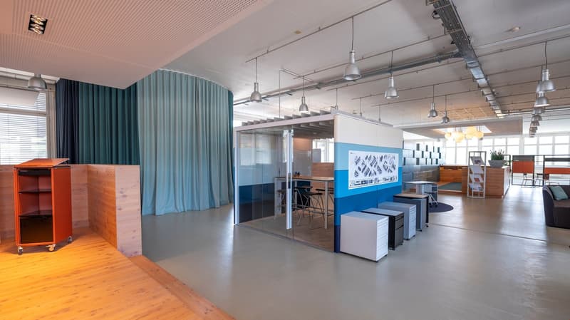 A bright and spacious office area with blue and white wall colors, multiple windows with blinds, a large partition with a glass door, several chairs, a wooden table, multiple cabinets, a couch with a pillow.