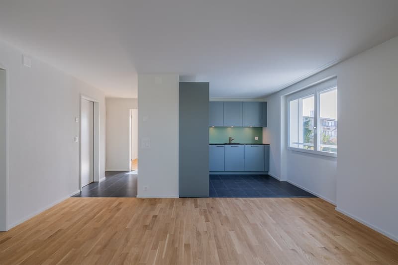 Empty room with wooden floor, white walls, kitchen with blue cabinets, sink, and window
