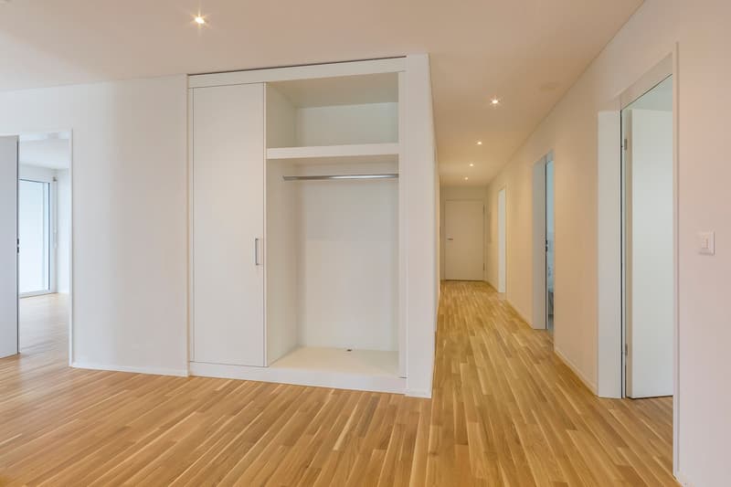Empty room with wooden floor, white walls, large wardrobe, and open doors leading to other rooms.
