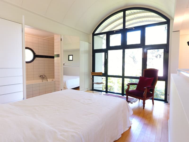 master bedroom with a large bed, wooden floor, bathtub, large arched window, chair